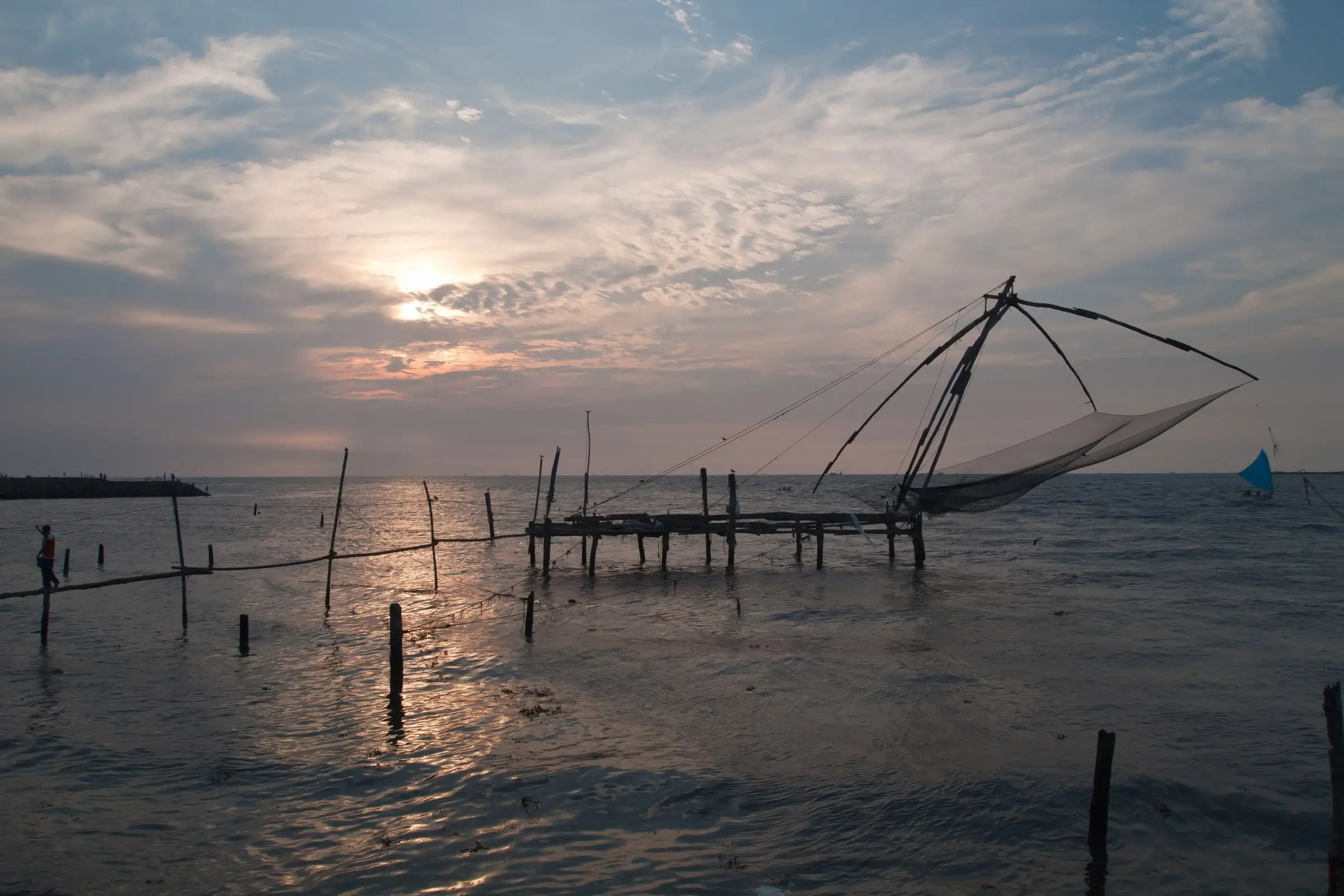 A Chinese fishing net on its wooden jetty silhouetted against a sun-lit evening sky over the sea.