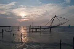 A Chinese fishing net on its wooden jetty silhouetted against a sun-lit evening sky over the sea.
