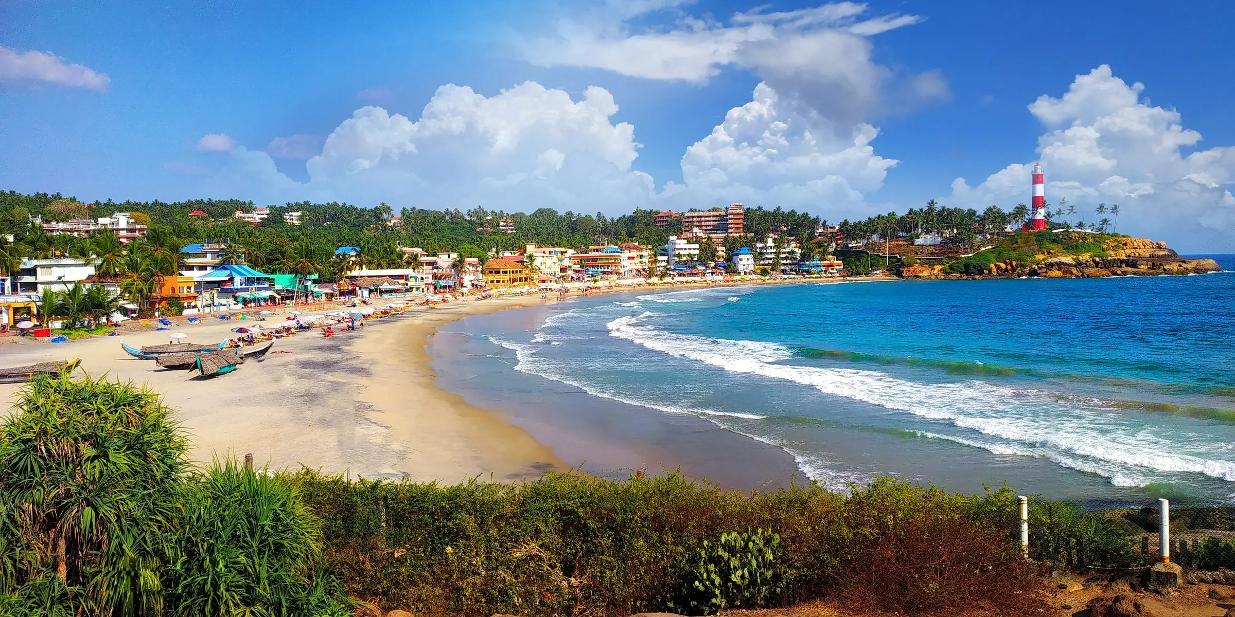 The crescent of Kovalam beach with colourful beachfront buildings, blue sea, and the red-and-white lighthouse on the headland at right.