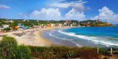 The crescent of Kovalam beach with colourful beachfront buildings, blue sea, and the red-and-white lighthouse on the headland at right.