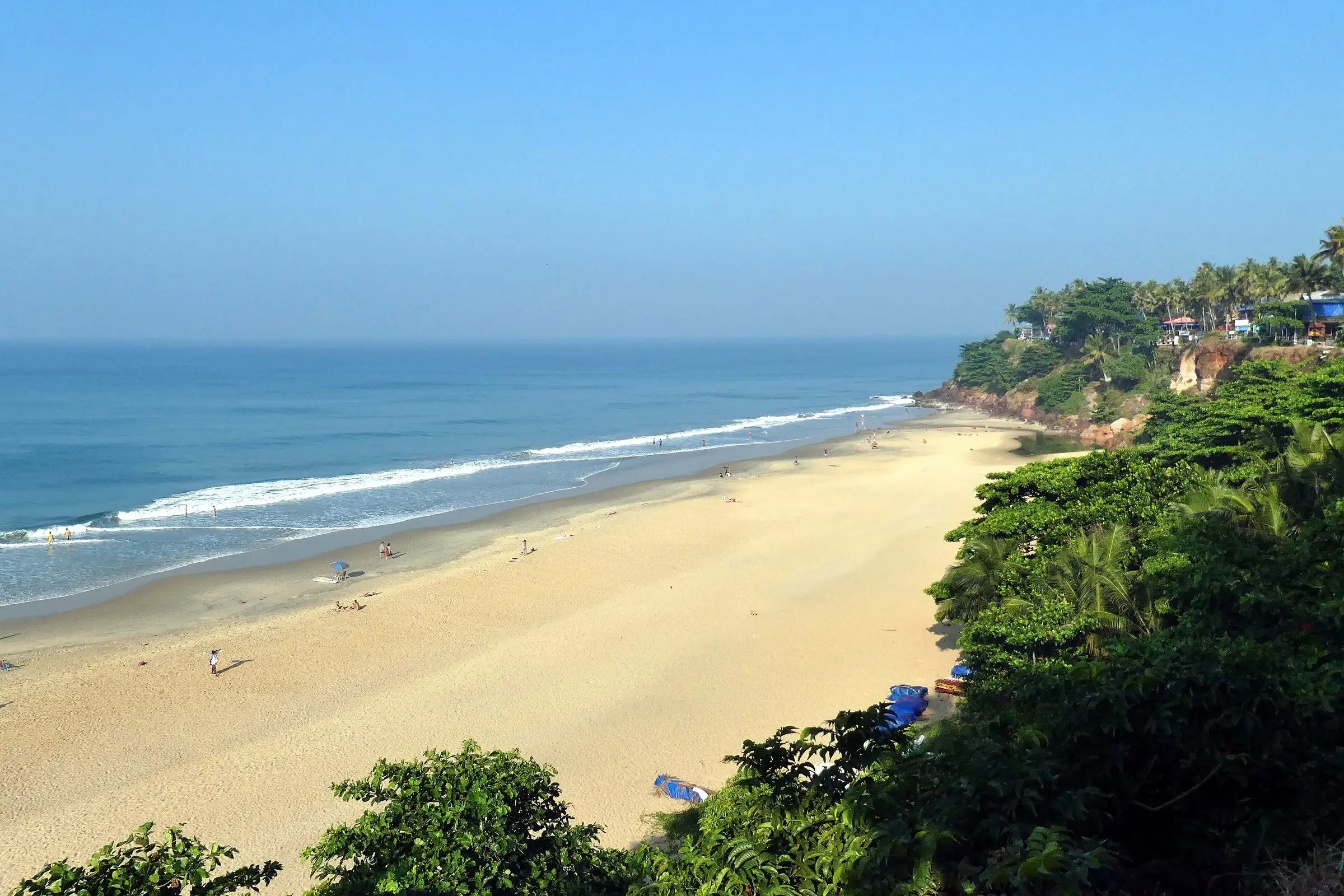 A long sandy beach seen from above, with blue surf on the left and a green cliff headland with palms on the right.